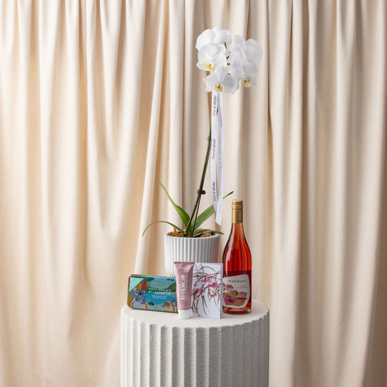 White orchid plant, bottle of pink wine, and small decorative items on a white pedestal against a beige curtain.