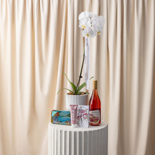 White orchid plant, bottle of pink wine, and small decorative items on a white pedestal against a beige curtain.