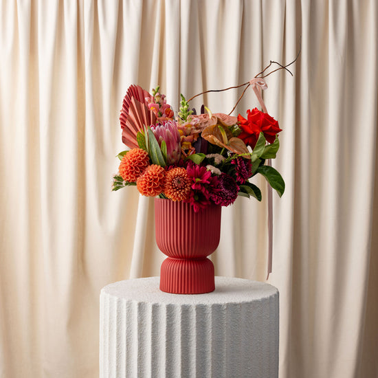 Colorful floral arrangement in a red vase on a white pedestal against a beige curtain background
