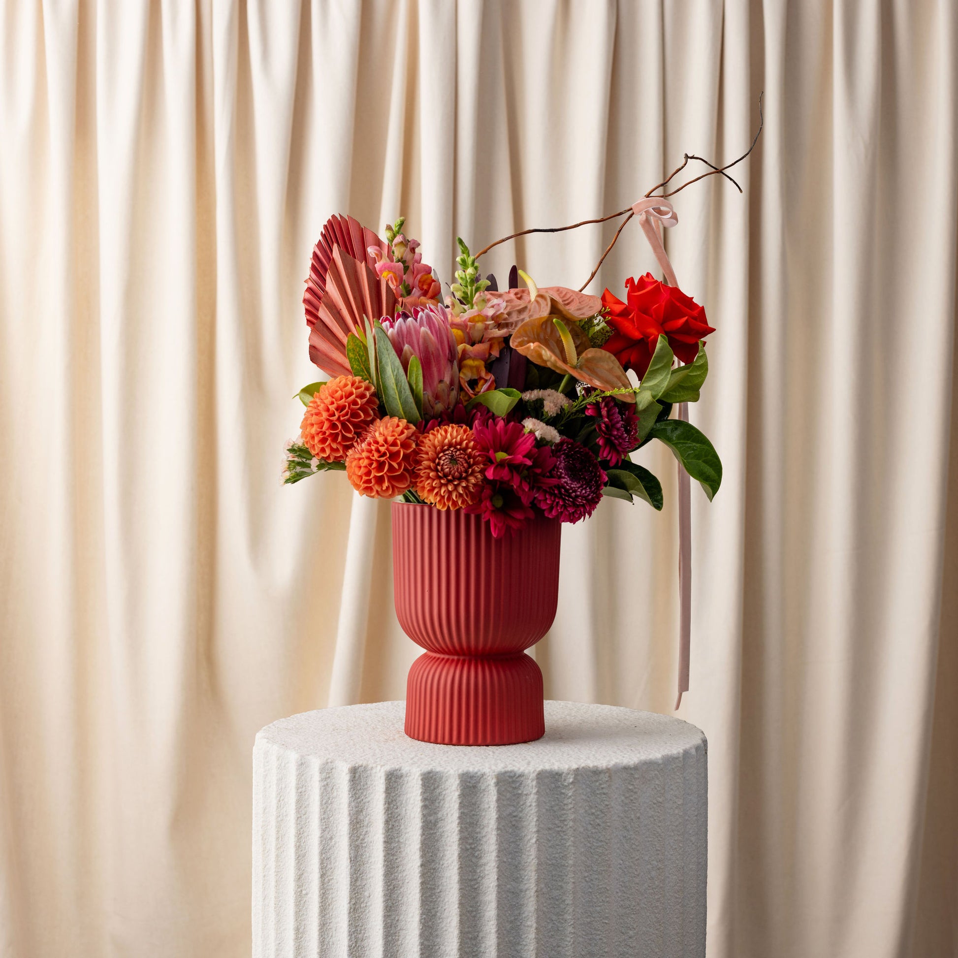 Colorful floral arrangement in a red vase on a white pedestal against a beige curtain background