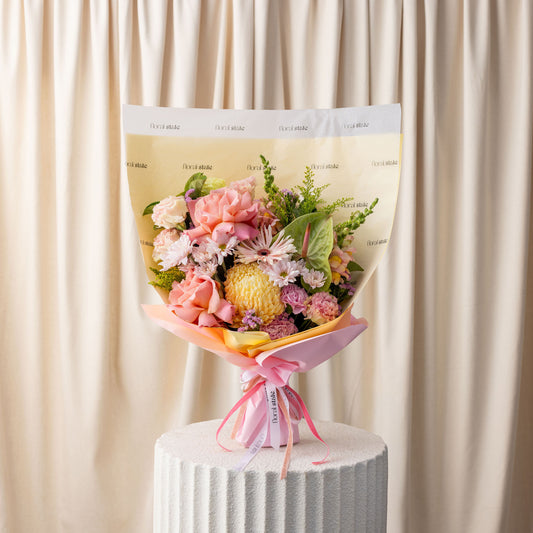 Bouquet of flowers wrapped in cellophane with a pink ribbon on a white pedestal against a beige curtain.