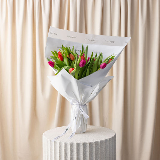 Bouquet of tulips wrapped in white paper on a cylindrical white stand with a beige curtain background