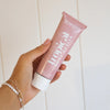 Hand holding a pink tube of tropical body butter against a white background