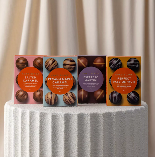 Four chocolate packaging boxes on a white pedestal with a beige curtain background
