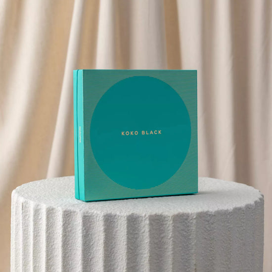 Teal box with 'Koko Black' branding on a textured white surface with beige curtains in the background