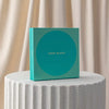 Teal box with 'Koko Black' branding on a textured white surface with beige curtains in the background