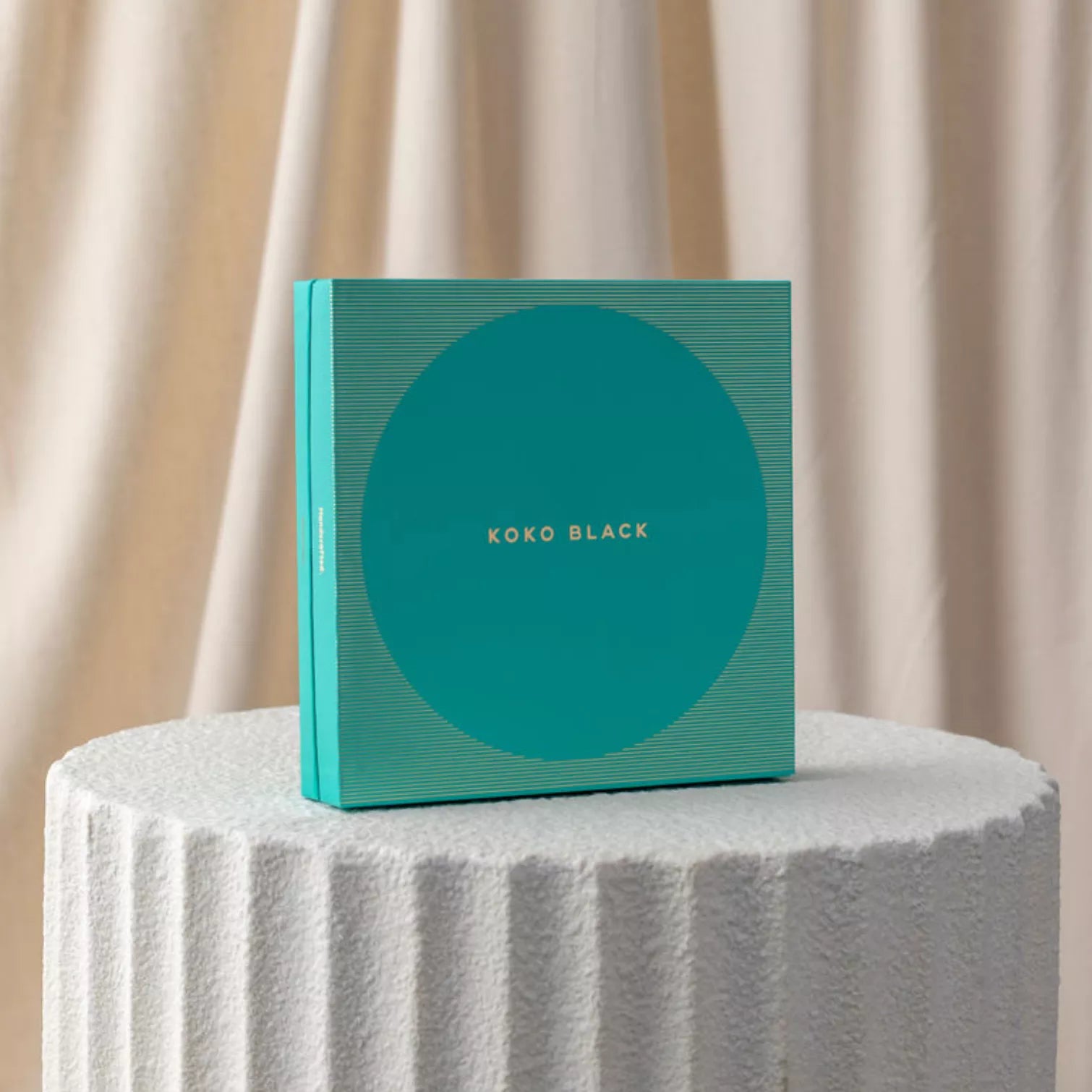 Teal box with 'Koko Black' branding on a textured white surface with beige curtains in the background